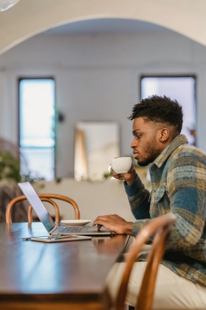 Focused young black man working remotely at home, sipping coffee, using a laptop.